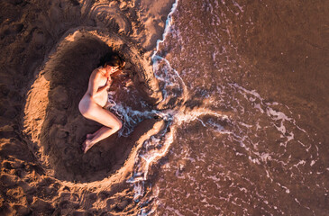 Mujer embarazada tumbada en la arena simulando un útero y el cordón umbilical.  - obrazy, fototapety, plakaty