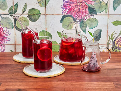 Raspberry ice tea in a special glass teapot, with two ice tea glasses and borosilicate straws, on a walnut table and ceramic tiles in the background.