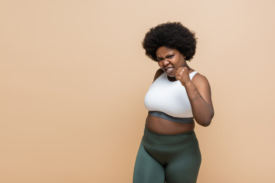 Emotional African American Plus Size Woman In Crop Top Showing Clenched Fist Isolated On Beige.