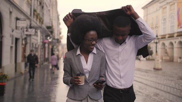 Happy afro american man and woman hiding from rain under jacket while walking on city street. Two business partners going to business meeting.