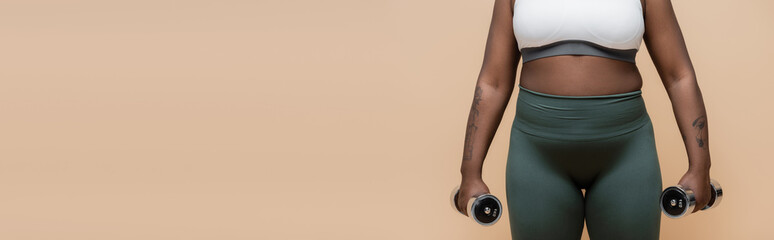cropped view of tattooed african american plus size woman in sportswear holding dumbbells isolated on beige, banner.