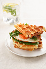 Savory breakfast - fresh homemade belgian waffle sandwich with green lettuce, crisp bacon and fried egg on white plate on beige colored table cloth, glass with lemon water