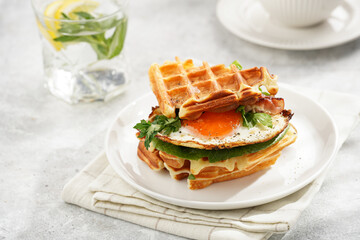 Savory breakfast - fresh homemade belgian waffle sandwich with green lettuce, crisp bacon and fried egg on white plate on beige colored table cloth, glass with lemon water, cup of coffee
