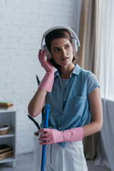 brunette woman in rubber gloves and wireless headphones looking away at home.