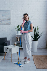 young woman adjusting wireless headphones while standing with mop in living room.