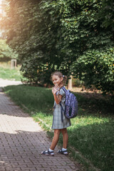 Fototapeta premium A little girl in a dress with pigtails holds large blue backpack, waving hand and walks on road alley. First september day in elementary school.