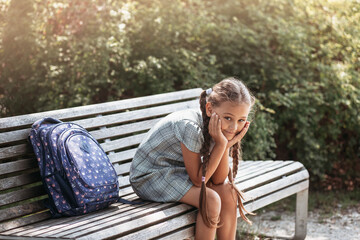 Back to school. A cute little schoolgirl in a dress with pigtails and large blue backpackis sitting on a bench in the school yard . A little girl is going to the first grade. - obrazy, fototapety, plakaty