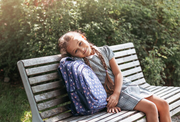 Back to school. A cute little schoolgirl in a dress with pigtails and large blue backpackis sitting on a bench in the school yard . A little girl is going to the first grade.