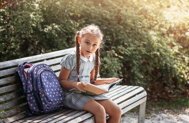Obraz premium Girl with a backpack sitting on a bench and reading a book near the school. Back to school, lesson schedule, a diary with grades. Education, primary school classes, Back to school