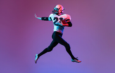 Fototapeta na wymiar Portrait of sportive man, american football player in uniform playing, training isolated over purple background in neon light.