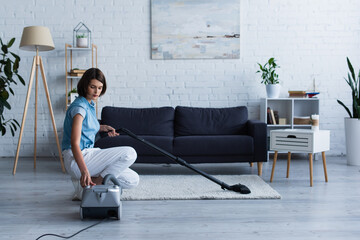 Brunette woman switching vacuum cleaner near carpet in living room.