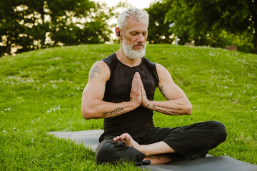 Mature gray sportsman wearing earphones meditating on yoga mat in park