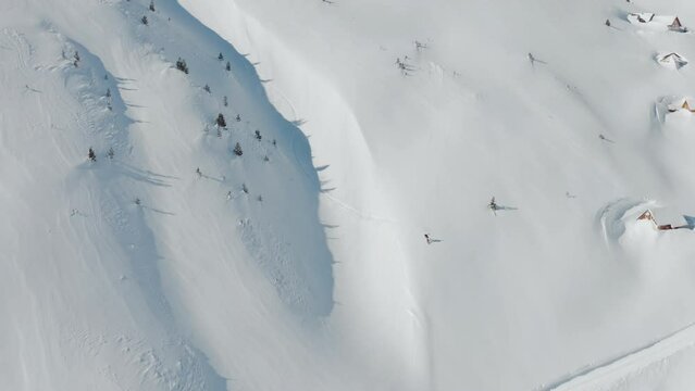 Aerial view of a freestyle snowboarder on a high-altitude mountain summit on a sunny winter day. He does spin tricks while riding off-trail fresh powder. Heliboarding remote pristine slopes.
