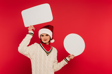 smiling young woman in santa hat and sweater holding speech bubbles isolated on red.