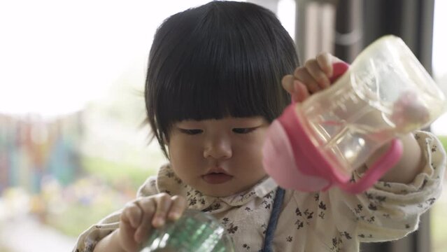 closeup view of an innocent female toddler kid playing with her sippy bottle and cup while drinking water in a restaurant at daytime.