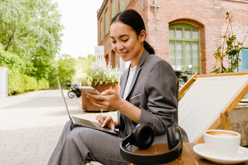 Asian woman smiling while using laptop and cellphone in cafe outdoors