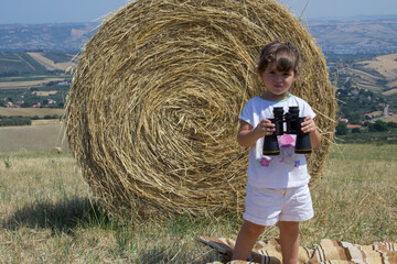 Naklejka na ściany i meble Picture of an adorable little girl playing watching views with binoculars against a background of straw bales in the middle of a field. 