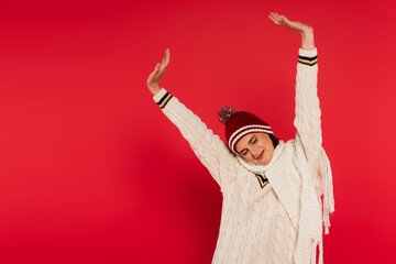 Cheerful woman in knitted clothes stretching on red background.