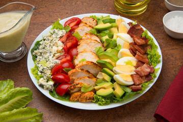 Cobb salad of romain lettuce, slices bacon, avocado, chicken, tomato, eggs, blue cheese in a white salad bowl on a brown background. - 527004611