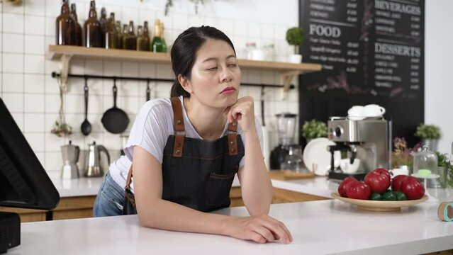 portrait of a bored asian waitress tapping finger and propping head on the counter while waiting for clients to visit the restaurant during pandemic outbreak