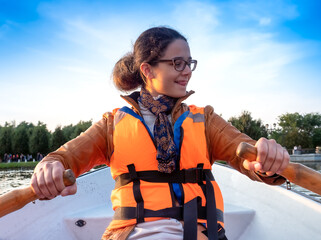Beautiful brunette woman teen with glasses and in an orange life jacket rowing oars while sitting in a boat. Family walks at the boat station. Illuminated by sun glare