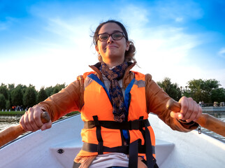 Beautiful brunette woman teen with glasses and in an orange life jacket rowing oars while sitting in a boat. Family walks at the boat station. Illuminated by sun glare