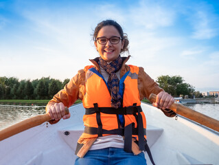 Beautiful brunette woman teen with glasses and in an orange life jacket rowing oars while sitting...