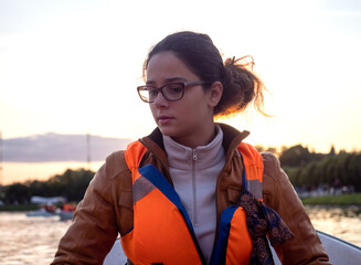 Beautiful brunette woman teen with glasses and in an orange life jacket rowing oars while sitting in a boat. Family walks at the boat station. Illuminated by sun glare