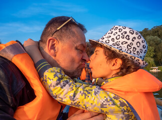 Father gives his son a kiss in a boat on the lake. Family in vests have fun during autumn walk in the park