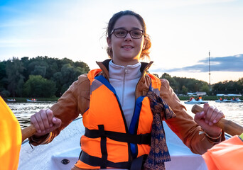 Beautiful brunette woman teen with glasses and in an orange life jacket rowing oars while sitting...