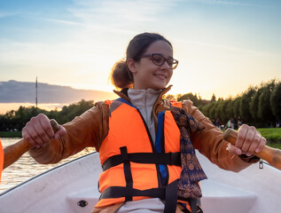 Beautiful brunette woman teen with glasses and in an orange life jacket rowing oars while sitting in a boat. Family walks at the boat station. Illuminated by sun glare