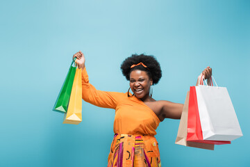 excited african american body positive woman holding shopping bags isolated on blue.
