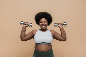 Fototapeta na wymiar happy african american plus size woman in sportswear exercising with dumbbells isolated on beige.