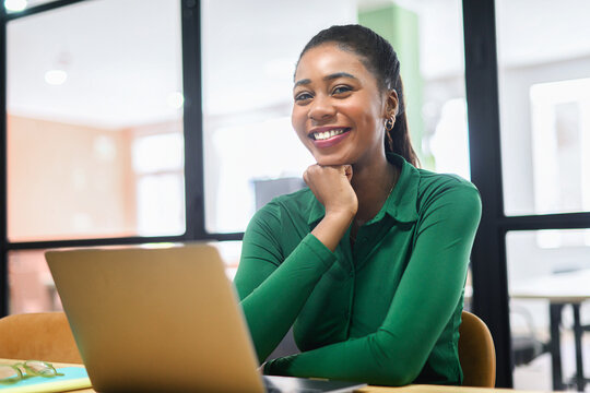 Happy Inspired African-American Female Office Employee Using Laptop Indoors, Resting Chin With Hand And Looking At Camera, Smiles. Excited Freelancer Woman Enjoys Remote Work In Coworking Space