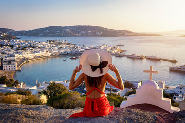 A elegant tourist woman in a red dress enjoys the view over the city of Mykonos island during a golden summer sunset, Cyclades, Greece - obrazy, fototapety, plakaty
