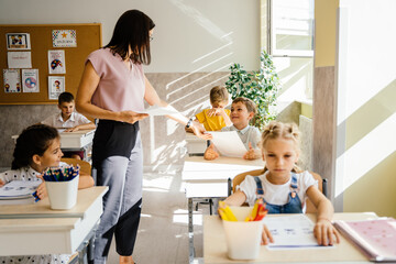 Primary school children interactive with the teacher. Female teacher distributes worksheets with tests for work in the lesson. Education and school concept. - obrazy, fototapety, plakaty