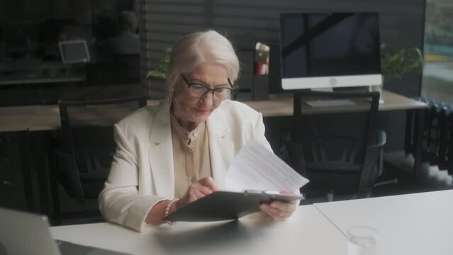 Serious older lawyer businesswoman in glasses reading signing trust partnership contract sit at table in office. Executive ceo put signature make legal bank sale financial loan investment agreement.