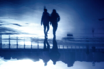 Silhouette of a couple reflected in the water of the street walking on a cold rainy winter day in...