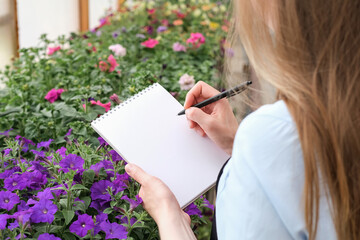 Young woman making notes in a notebook while working in an industrial greenhouse. Close-up of female hands with an empty notebook and pen. Notepad mockup with place for text