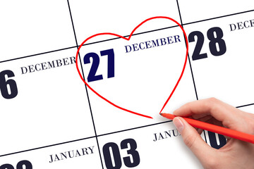 A woman's hand drawing a red heart shape on the calendar date of 27 December. Heart as a symbol of...