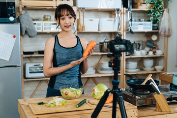 asian athletic woman holding a carrot and talking to digital camera on tripod is showing the...