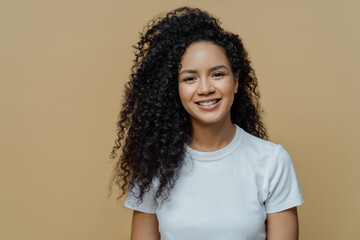 Horizontal shot of curly haired woman with healthy skin, toothy smile, enjoys being photographing, wears casual white t shirt, isolated on beige background. People, emotions and lifestyle concept