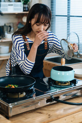 vertical closeup shot of an Asian woman holding the lid is covering her nose with disgust face after a bad smell from the soup pot on gas stove in the kitchen.