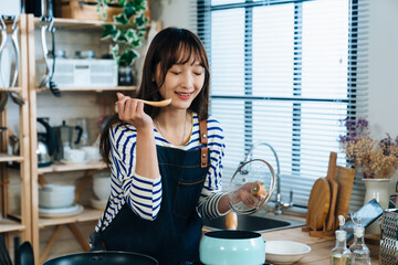 happy Asian housewife is smiling with satisfaction while trying soup from the spoon over the pot at...