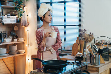 asian woman wearing pajamas with hair wrapped in a towel is looking outside kitchen window in contemplation while having tea on a tranquil night at home.