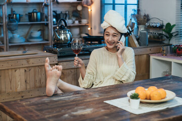 carefree Asian girl with towel on head and legs on the dining table is enjoying wine while chatting happily on phone after shower at night in cozy home interior