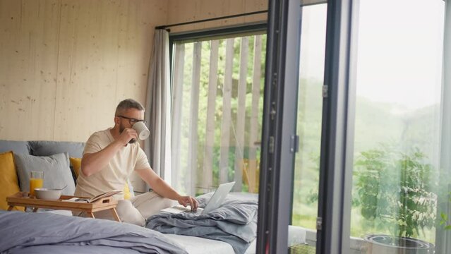 Mature man having breakfast and working in tiny house, in front of nature. Freelancer office concept