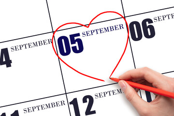 A woman's hand drawing a red heart shape on the calendar date of 5 September . Heart as a symbol of...