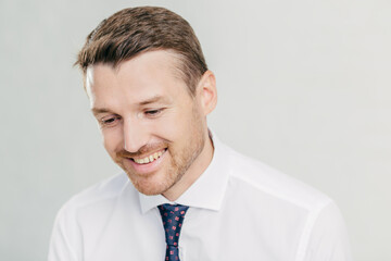 Joyful male corporate worker in elegant white shirt, looks happily down, remembers something pleasant in mind, has toothy smile, wears elegant white shirt with tie, isolated over white wall.