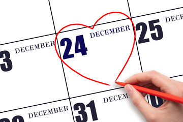 A woman's hand drawing a red heart shape on the calendar date of 24 December. Heart as a symbol of...